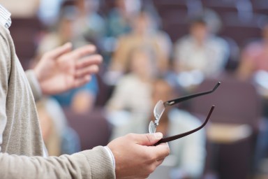 Professor holding eyeglasses and gesturing in lecture hall