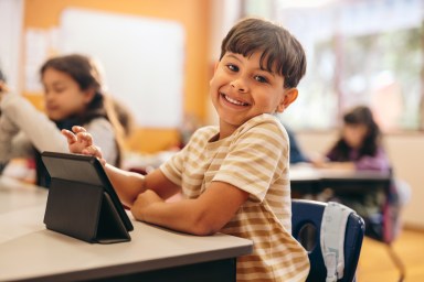 Child development in action as young kid learns to code with a tablet in school. Happy little boy looking at the camera while sitting at his desk in a digital literacy class. Male student in primary school.