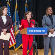 three people at a podium discussing misinformation during Election Day with flags in the back