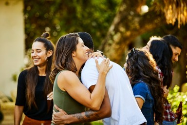 Happy woman embracing friend in backyard during a visit. Male and female hipsters enjoying weekend together. They are wearing casuals.