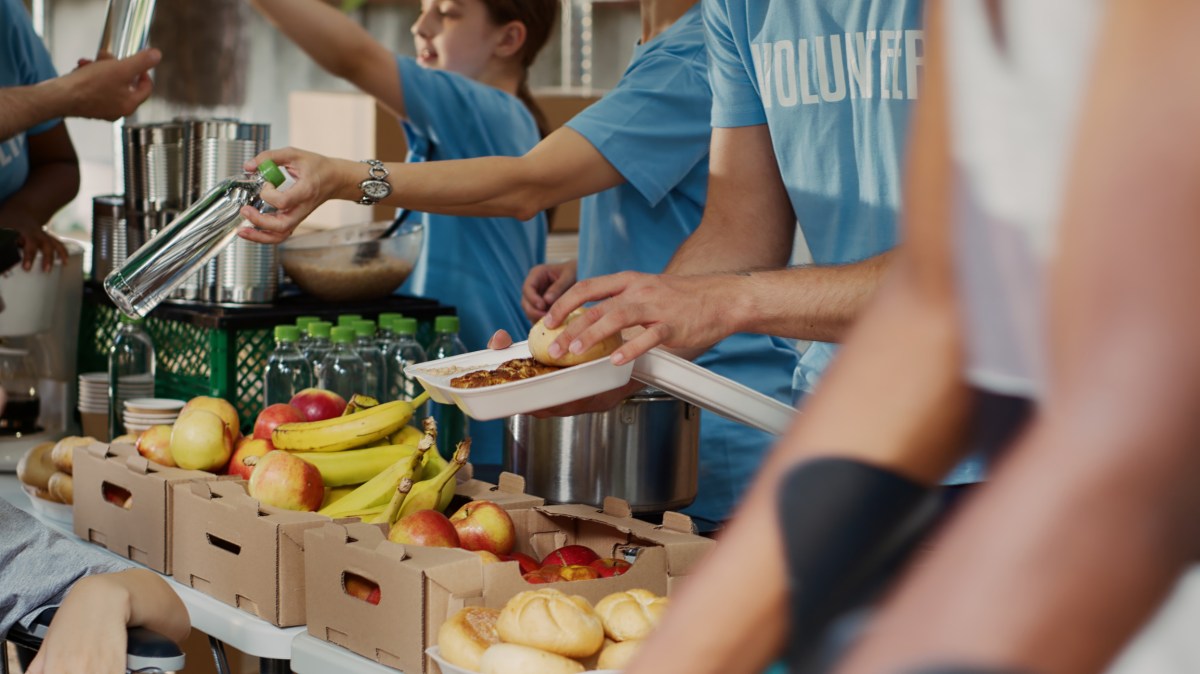 At food drive, poor female wheelchair user receives free food and provisions. Multiethnic volunteers in blue t-shirt distributing fresh fruits and hot meals to less privileged. Close-up, tripod.