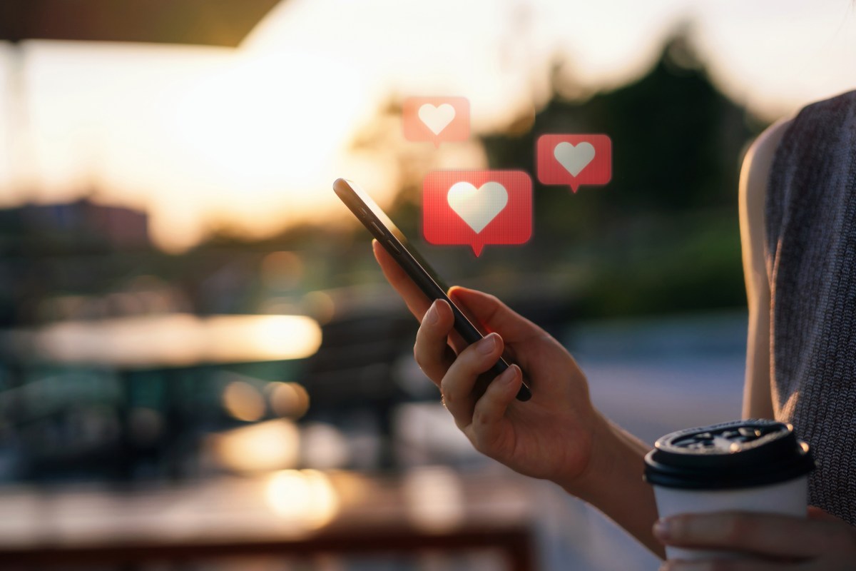 Close up of young woman checking social media network on smartphone on the go in the city. Mobile phone with heart emoji speech bubble message on the screen. People engaging in networking with technology. Social media addiction concept