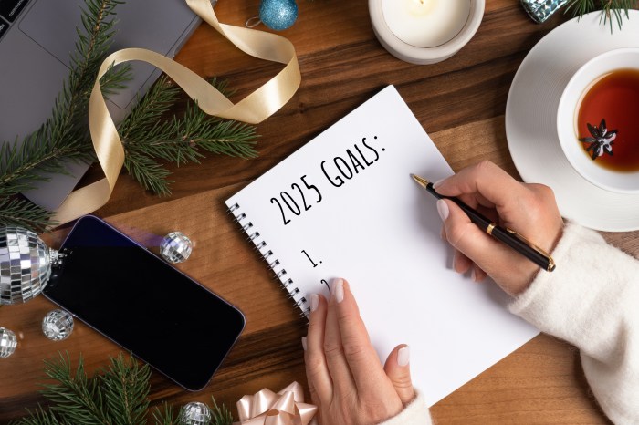 Concept of Writing the Goals on Merry Christmas and Happy New Year Holidays with woman's hands, hot tea cup, fir tree branches, candle and notebook with written 2025 Year goals on wooden table with Christmas decorations and computer. Woman writing New Year's goals. Top view, flat lay, copy space. Close-up. Minimalistic background with copy space.