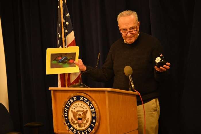 Sen. Chuck Schumer at a podium holding a magic 8 ball and photo of a drone