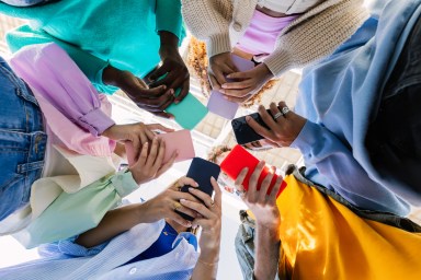 Low angle view of young group of people holding cellphone devices at city street. Diverse addicted teen friends watching social media content on smartphone app. Youth, gen tech people and technology lifestyle concept.