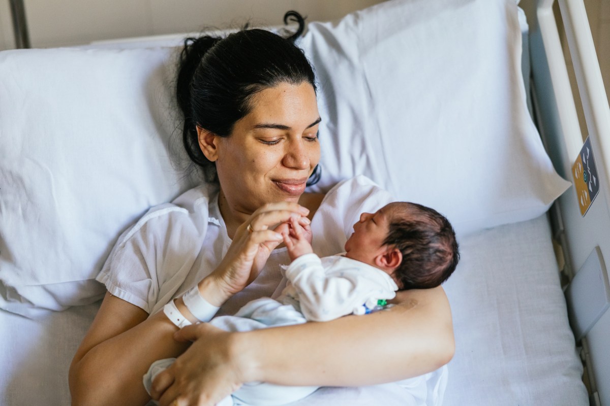 Woman holds the hands of her newborn baby as she rests in a hospital bed. She gazes lovingly at her tiny baby.