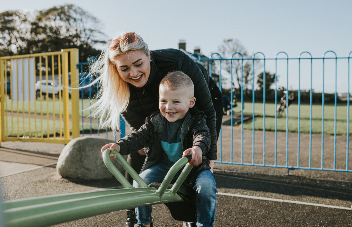 Mother holds her son while he rides on a green metal seesaw in a sunny park. They both smile. He grips the handles tightly.