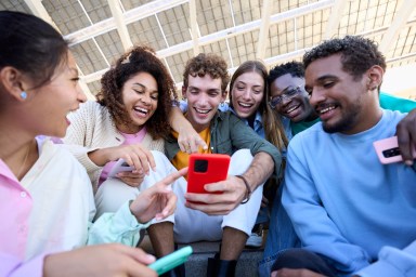 Mixed raced group of friends gathered together using a phone looking at social network content smiling and enjoying.