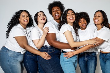 Group of females standing on white background and laughing. Multi-ethnic women in white t-shirt and blue jeans hugging each other.