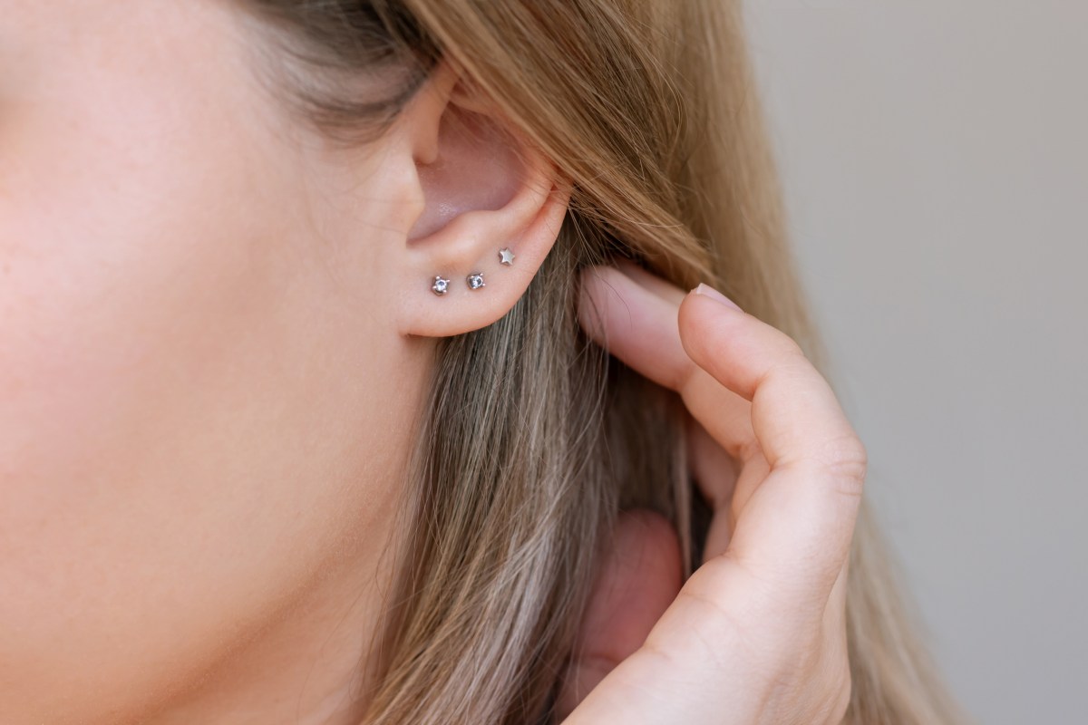 Ear piercing. Cropped shot of a young blonde woman wearing three stud earrings on the earlobe on a grey background. Jewelry with gemstones, accessories with fianits