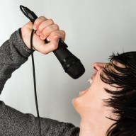 The young black hair guy is singing or screaming in the microphone on gray background.